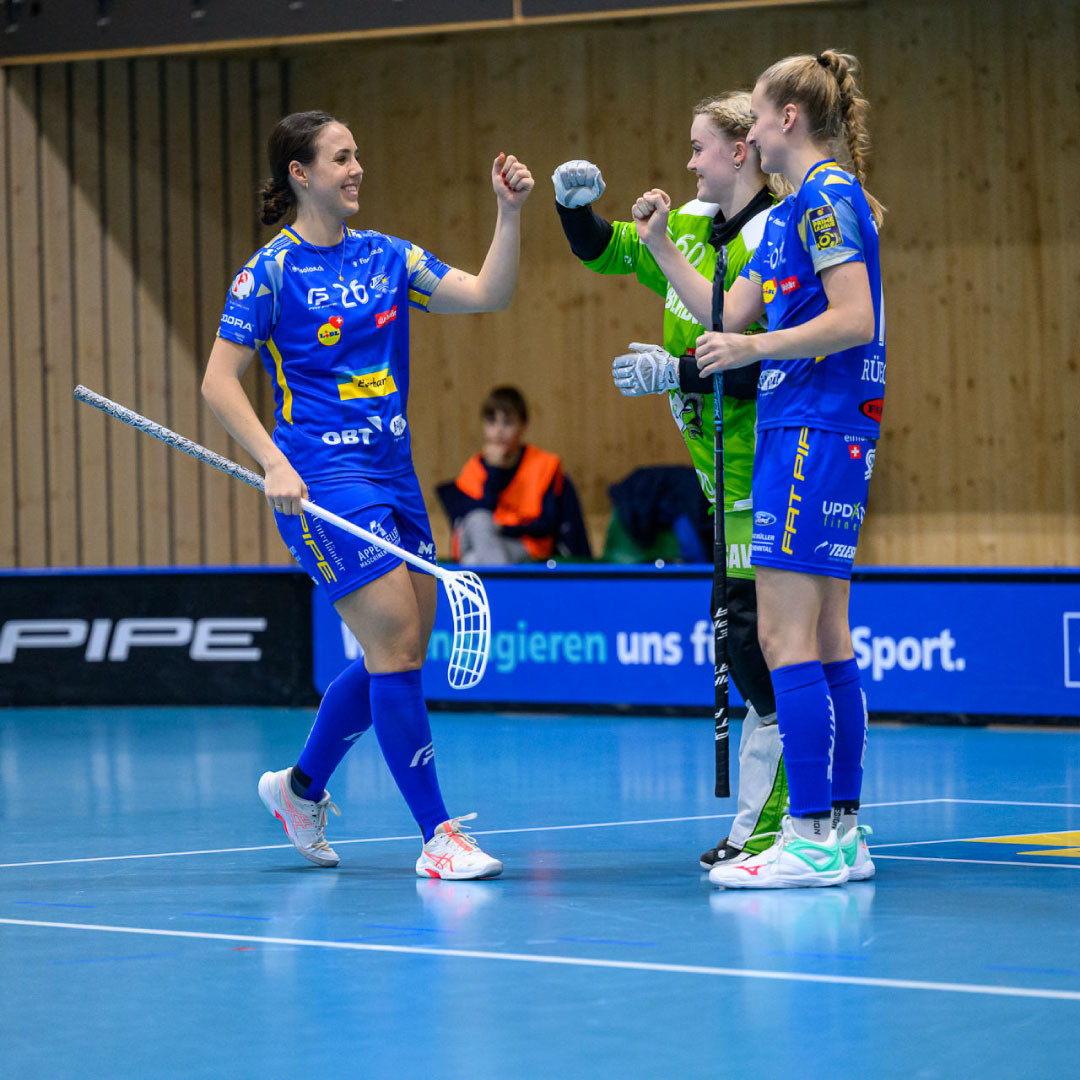 Celine Stettler in blauem Trikot schlägt lachend mit zwei Teamkolleginnen auf dem Unihockeyfeld ein