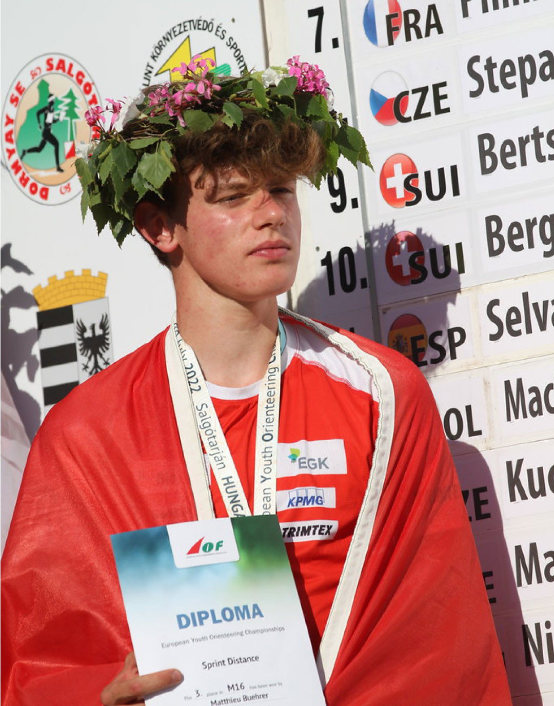 Matthieu Bührer mit Kranz auf dem Kopf und rotem Umhang hält Diplom in der Hand