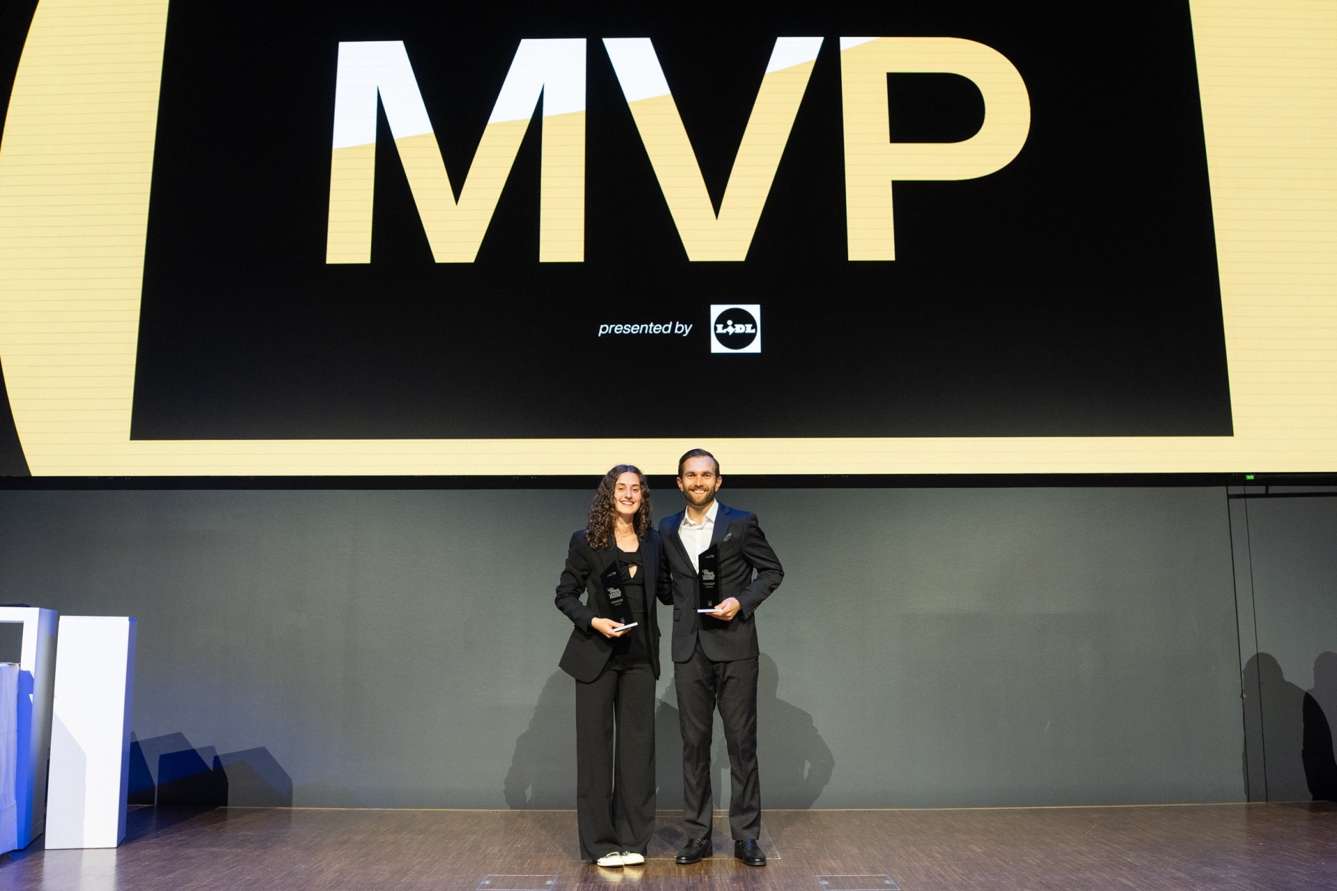 Marcia Wick pose devant la toile sur laquelle sont inscrites les lettres majuscules « MVP », en compagnie d'une autre personne